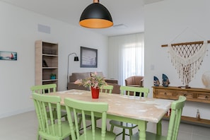 Uma sala de jantar com uma mesa, cadeiras, uma luminária pendente, uma estante, um quadro emoldurado e um enfeite de macramé na parede.