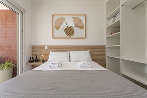 Cama bem arrumada com lençóis e toalhas brancas, cabeceira de madeira, obra de arte na parede com representação de leques e um armário de madeira com toalhas dobradas.