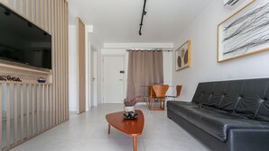 A modern living room with a flat-screen TV, a wooden coffee table, a black leather sofa, and a dining area with chairs. - Gastronomy | 120m from the Beach | Pool #CAMP07 (Florianópolis)
