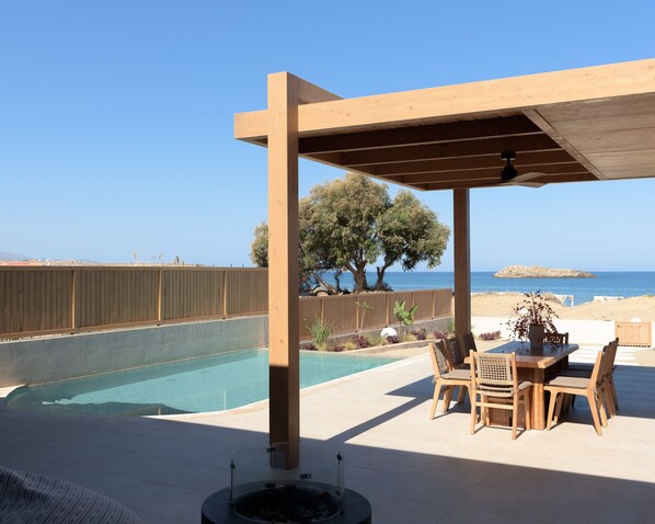 A covered outdoor area with a pool, wooden furniture, and a view of the sea.