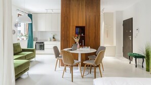A modern living space with a central wooden partition, a dining area with a round table and chairs, a green sofa, and a kitchen in the background.