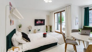 A modern hotel room with a bed, a desk with a laptop, a TV displaying 'NETFLIX', and a large window with curtains.