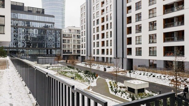 A modern residential building complex with multiple apartment buildings, a central courtyard with snow, and a tall skyscraper in the background.