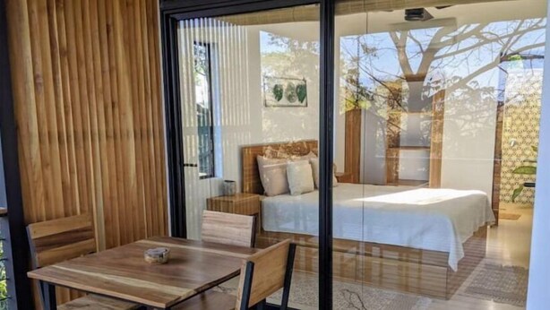 Ein modernes Schlafzimmer mit einem großen Schiebeglasfenster, das auf einen Balkon führt, einem Holztisch mit Stühlen und einem Bett mit weißer Bettdecke.