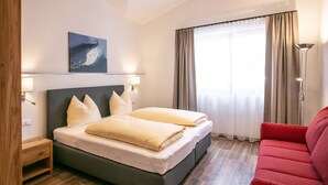 A hotel room with a bed, a red sofa, a painting on the wall, and a window with curtains. - Appart Hotel Amadeus  (Sankt Johann in Tirol)