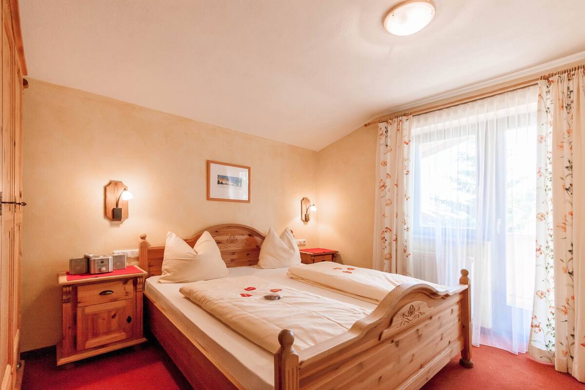 A bedroom with a wooden bed, two bedside tables, a nightstand, a wall-mounted lamp, and a window with sheer curtains.