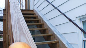 Un escalier en bois, doté d’une rampe, mène à un bâtiment blanc qui possède une fenêtre et une porte.