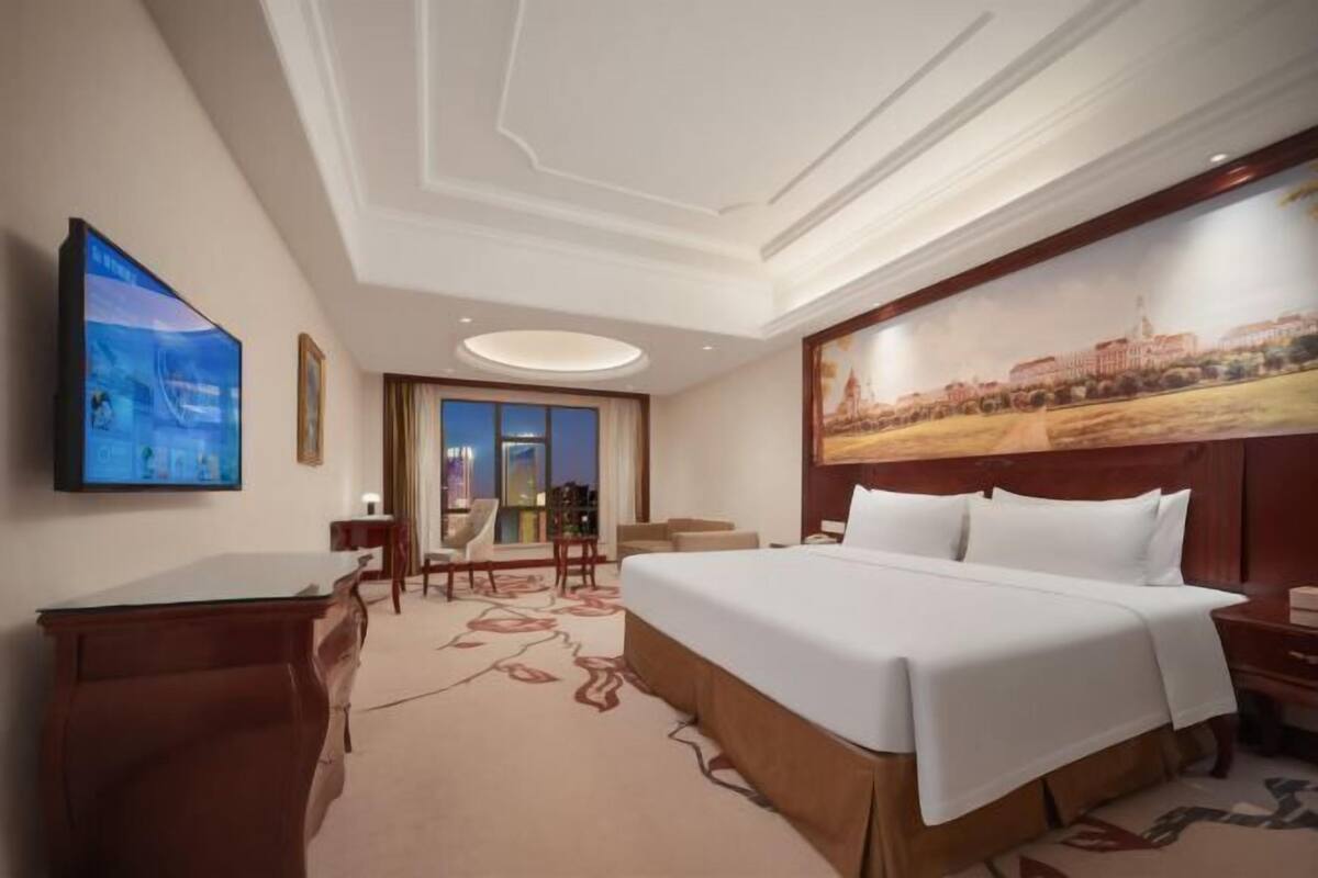 A hotel room with a large bed, a desk, a chair, a TV, and a painting on the wall.
