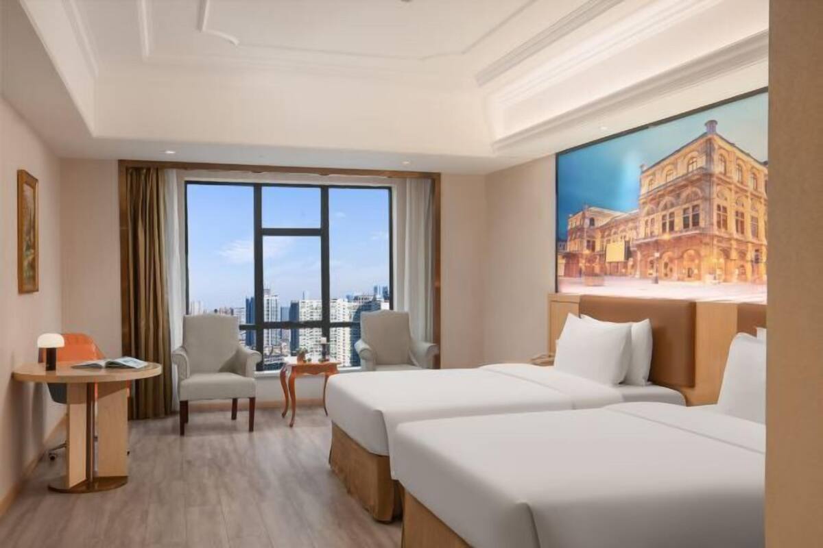 A hotel room with two beds, a desk, a chair, a view of the city, and a painting on the wall.