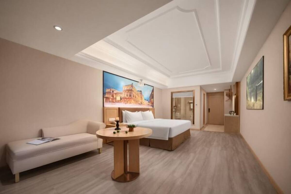 A hotel room with a bed, a sofa, a round table, and a large window with a cityscape view.
