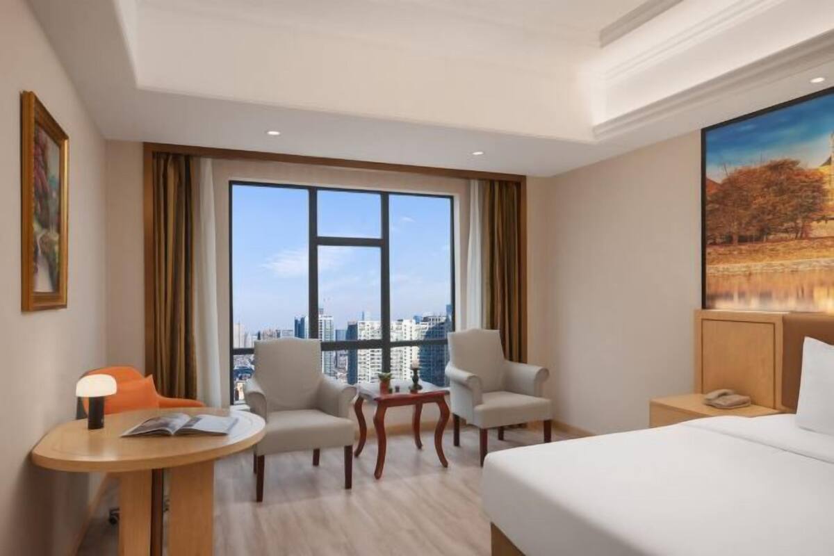 A hotel room with a large window, a bed, two armchairs, a small table, and a nightstand.