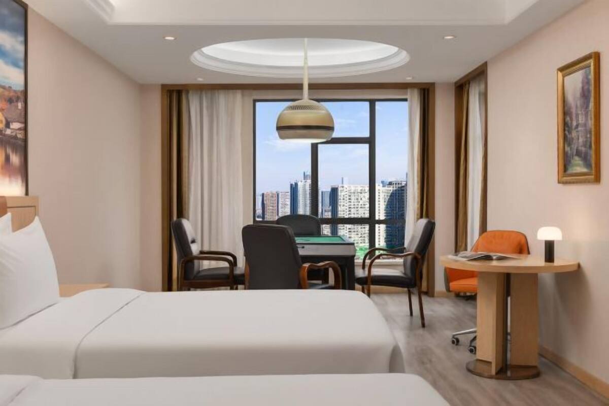 A hotel room with a bed, a desk with chairs, a view of the cityscape, and a large window.
