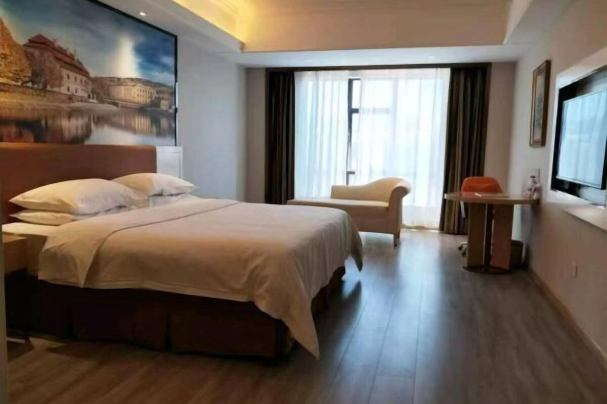 A hotel room with a bed, a chair, a desk with a computer, and a large window with curtains.