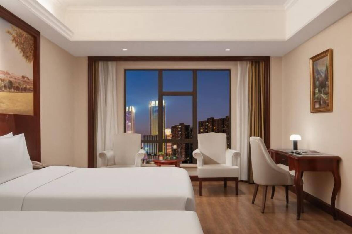 A hotel room with two beds, a desk, a chair, and a large window with a city view.