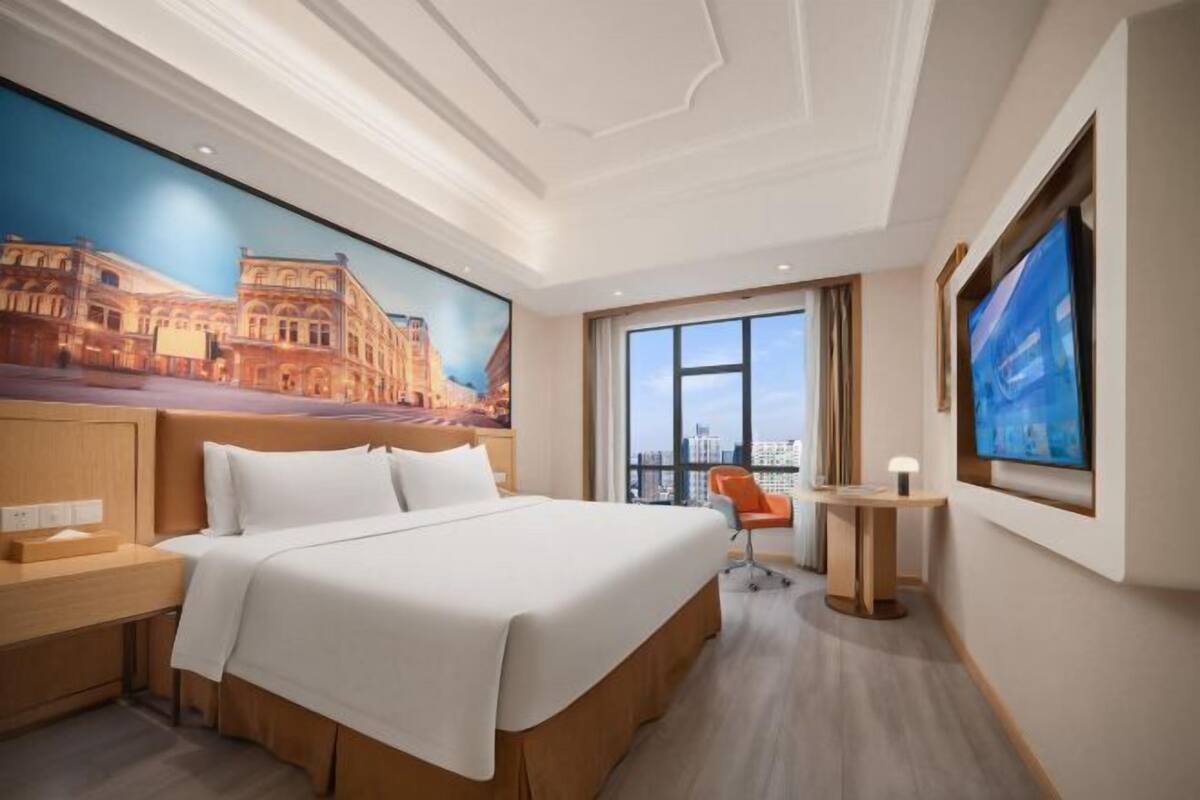 A hotel room with a large bed, a nightstand, a desk, a chair, a window with a city view, and a mural on the wall.