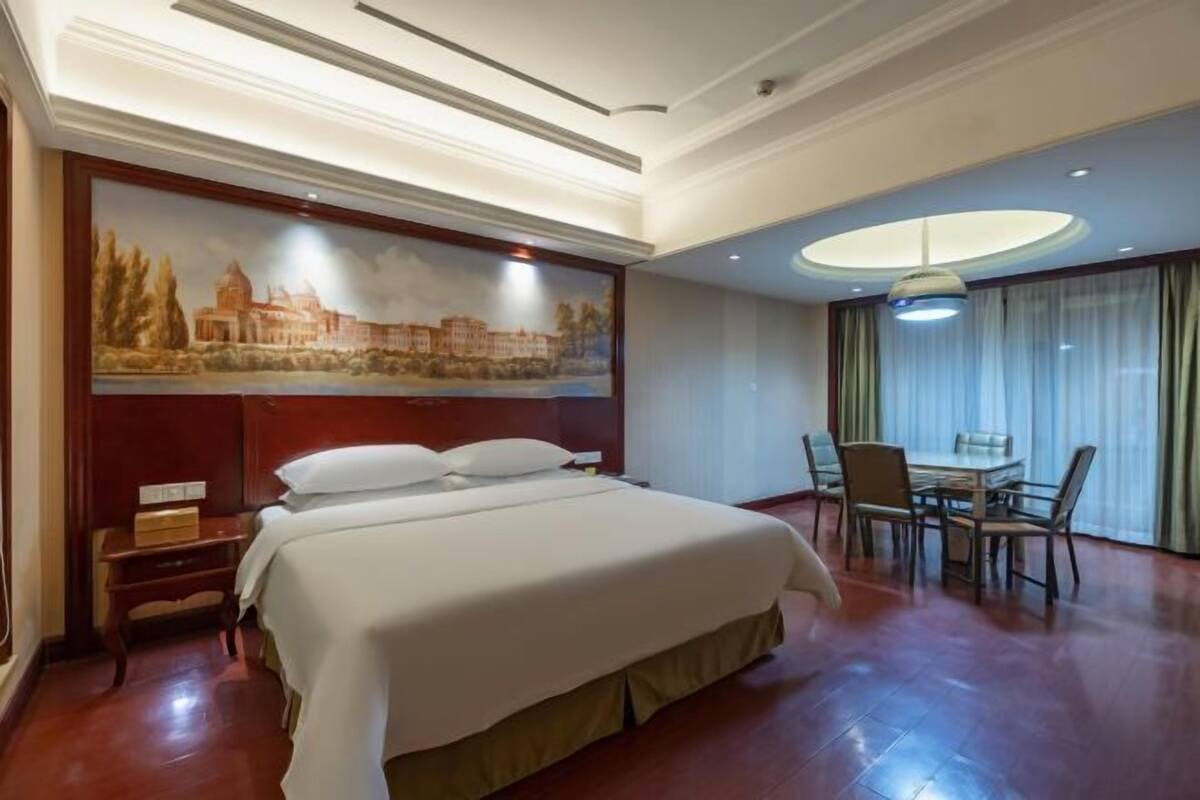 A hotel room with a large bed, a desk with a chair, a dining table with chairs, and a painting on the wall.