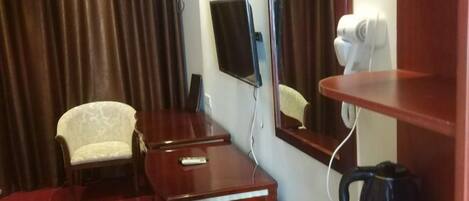 A hotel room with a wooden desk, a chair, a flat-screen TV, a mirror, a hairdryer, and a coffee maker.