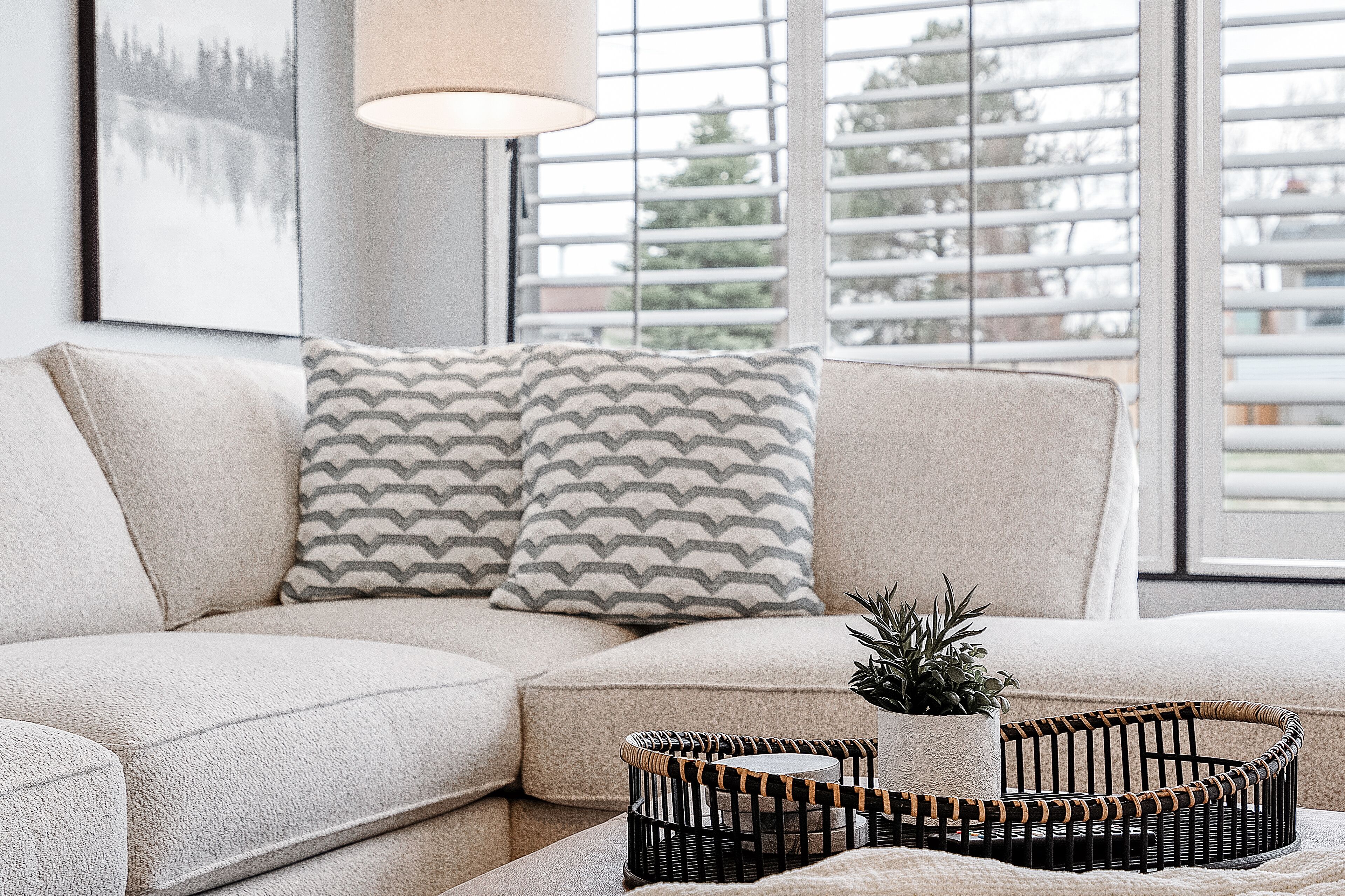 En moderne stue med en modulsofa, puder med mønster, et lille sofabord med en potteplante og et vindue med persienner.