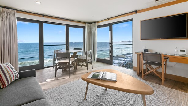 A modern hotel room with a large window overlooking the sea, a flat-screen TV, a desk, and a comfortable seating area.