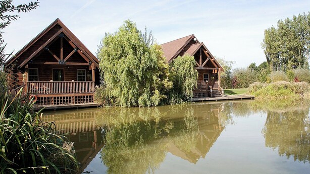 Two wooden cabins with balconies, a pond, and surrounding greenery.