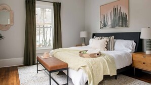 Un dormitorio con cama, mesitas de noche, un banco, una ventana con cortinas, un espejo en la pared y un cuadro.
