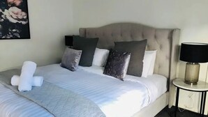 Un lit bien fait, avec une couverture et des oreillers gris, une lampe de chevet et une œuvre d’art florale encadrée accrochée au mur.