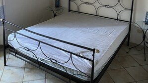 Un letto singolo con struttura in metallo e biancheria da letto bianca in una stanza con pavimento piastrellato e parete bianca.