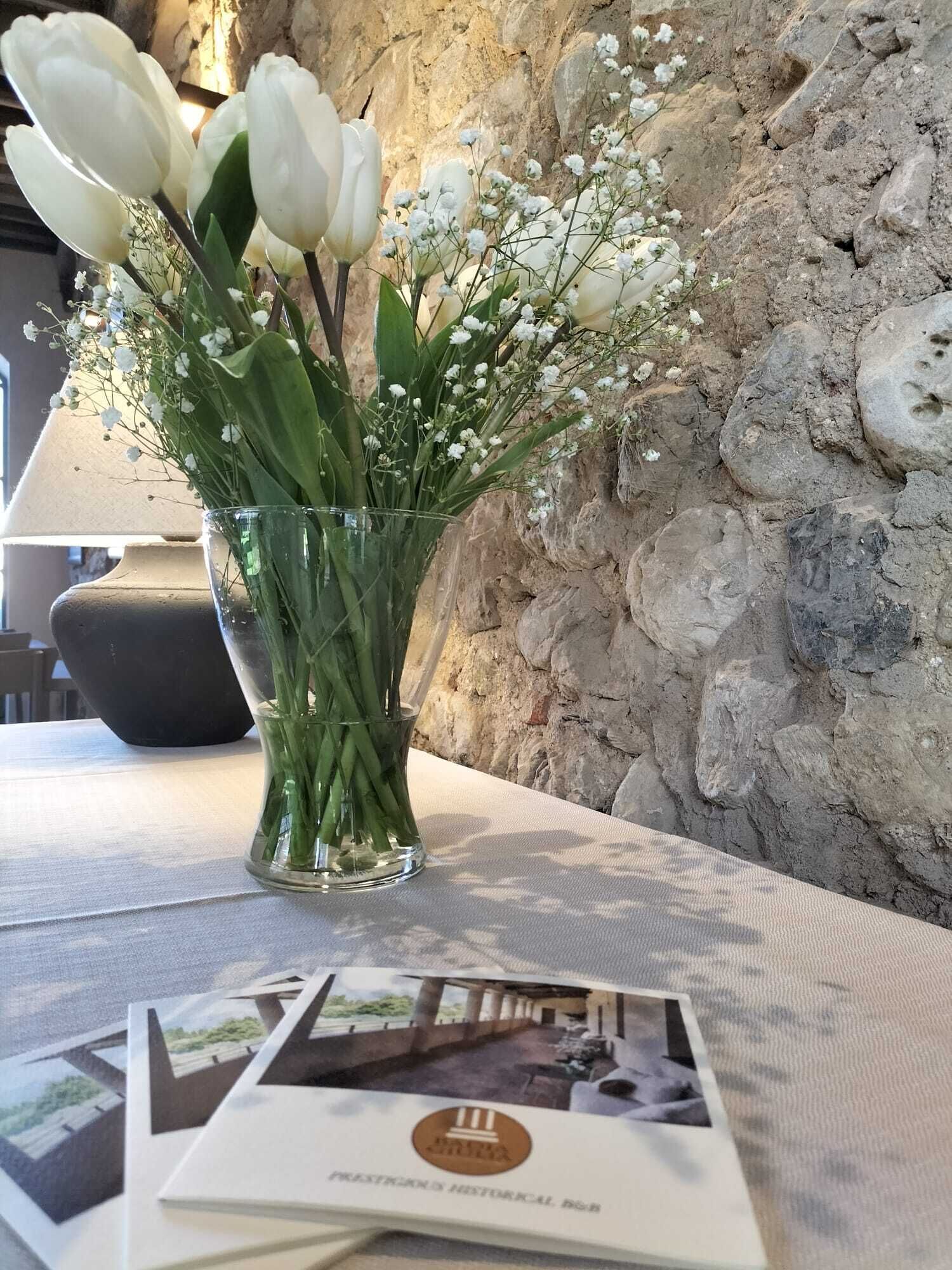 A clear glass vase with white tulips and baby's breath on a table against a stone wall.