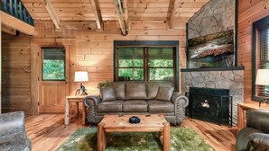 Interior acogedor de una cabaña de troncos con chimenea de piedra, muebles de madera y un ventanal con vistas a follaje.