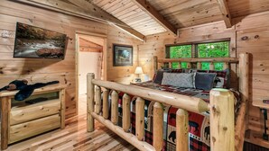 Habitación de cabaña de madera con una cama, un banco de madera, un televisor y una ventana con vistas a follaje.