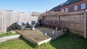 Een achtertuin met een houten terras, tuinmeubilair en een bakstenen gebouw op de achtergrond.