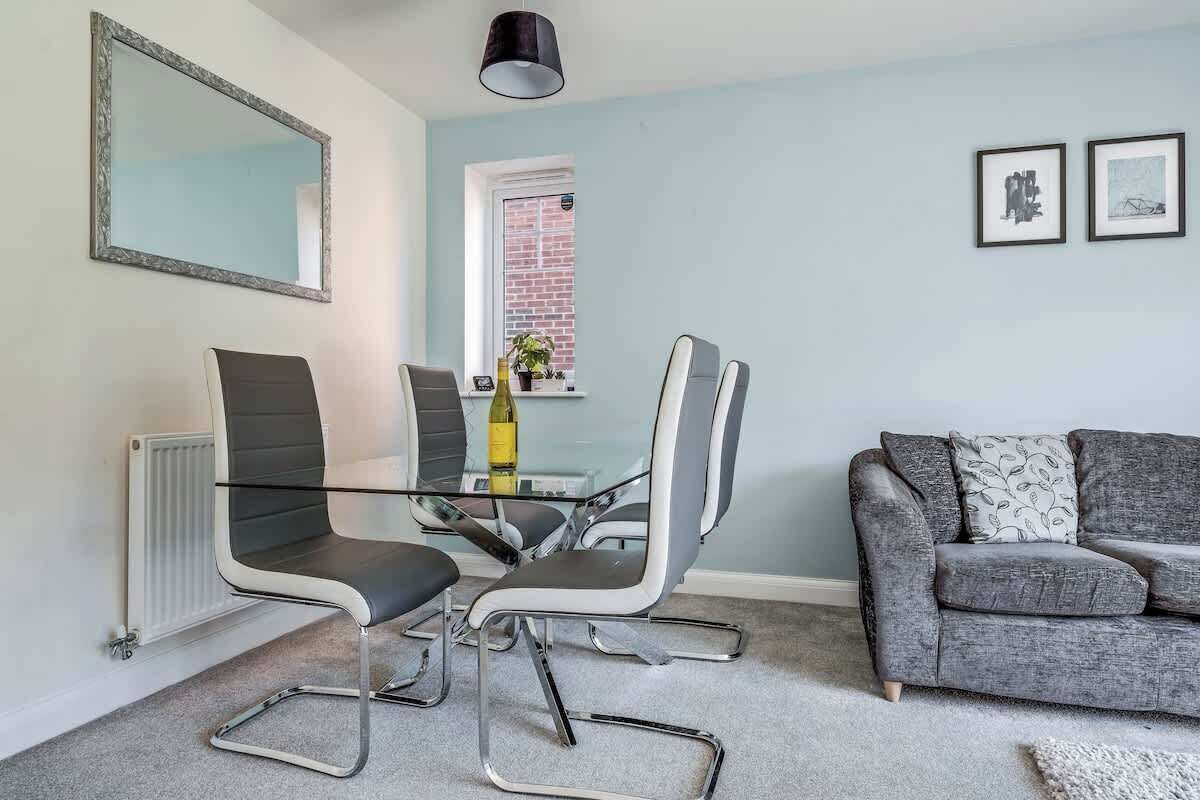 A modern living room with a grey sofa, a glass dining table with four chairs, a large mirror, and two framed pictures on the wall.