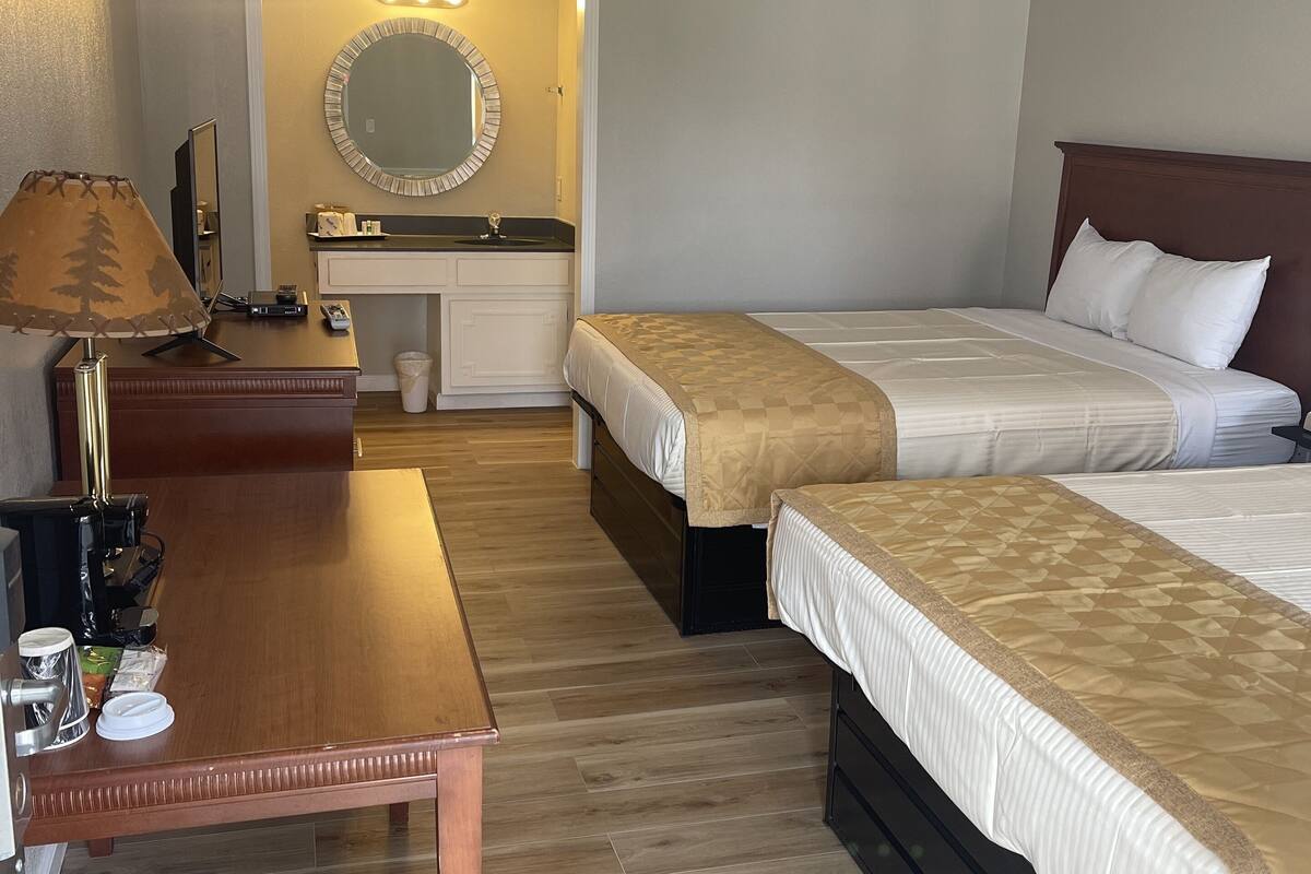 A hotel room with two single beds, a wooden desk with a lamp, a mirror, and a small vanity with a sink.