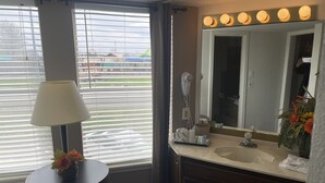 A hotel bathroom with a large window, a round table with a lamp, a mirror with lights, and a sink. - Branson Victorian Inn (Branson)