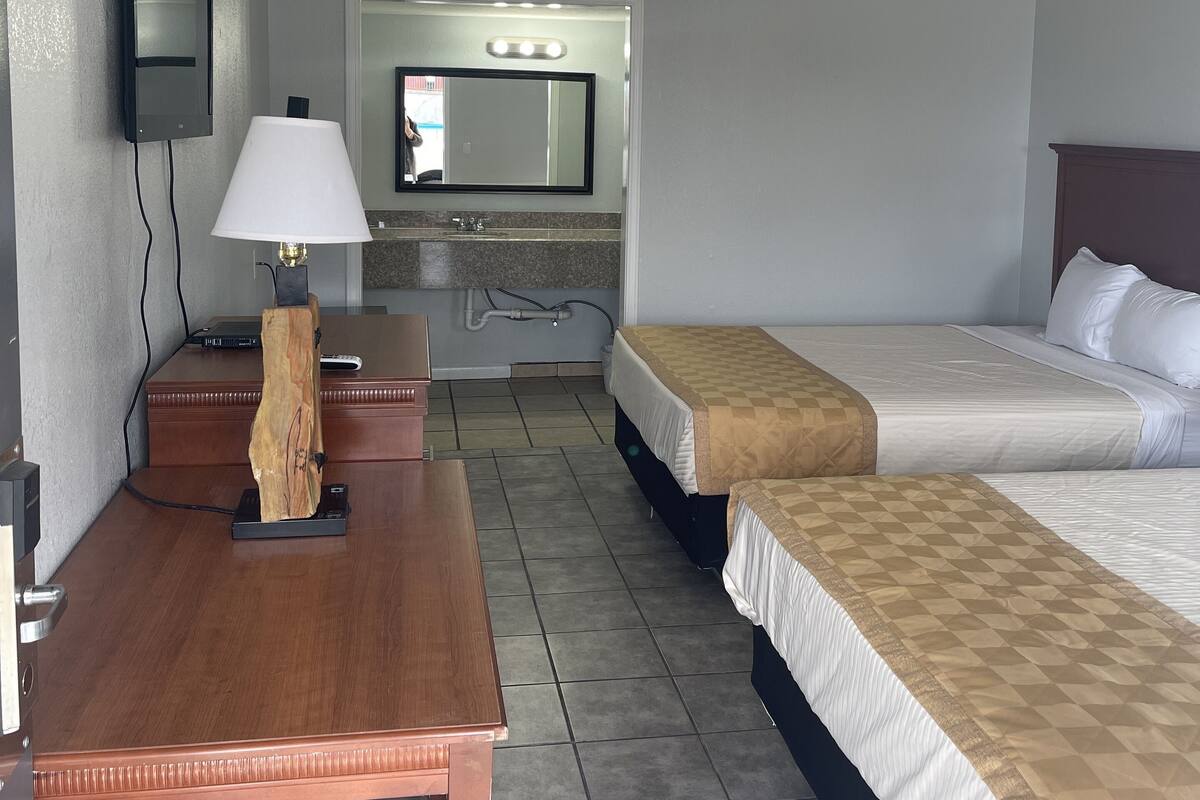A hotel room with a wooden desk, a lamp, a television mounted on the wall, a mirror above the mirror, and two beds with checkered bedspreads.
