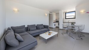 Un salon moderne avec un canapé d’angle gris, une table basse blanche et une table à manger en verre, accompagnée de chaises.