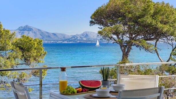 Un balcon avec une table préparée pour le petit-déjeuner, offrant une vue sur la côte, avec un voilier et des montagnes en toile de fond.