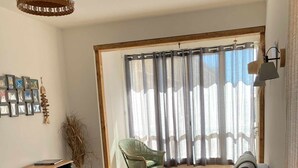 A living room with a sofa, a chair, a small table, and a cabinet. There is a large window with curtains, a hanging light fixture, and a wall-mounted picture frame.