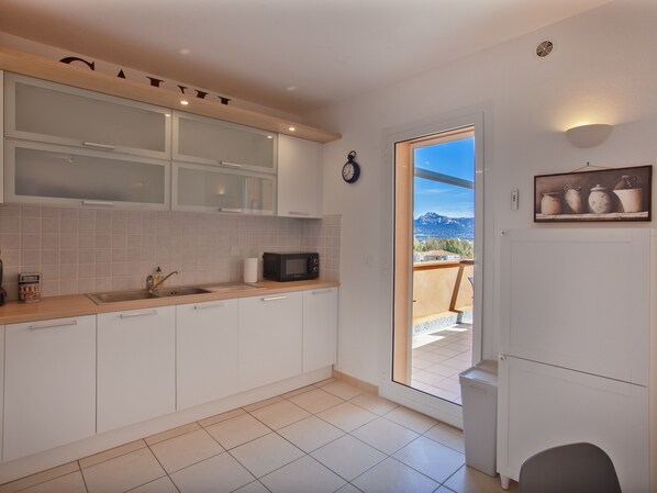 A modern kitchen with white cabinets, a microwave, and a sink. There's a door leading to a balcony with a view of mountains. - casa notte : appart spacieux, calme, Plage et centre de calvi 15 min à pied (calvi)