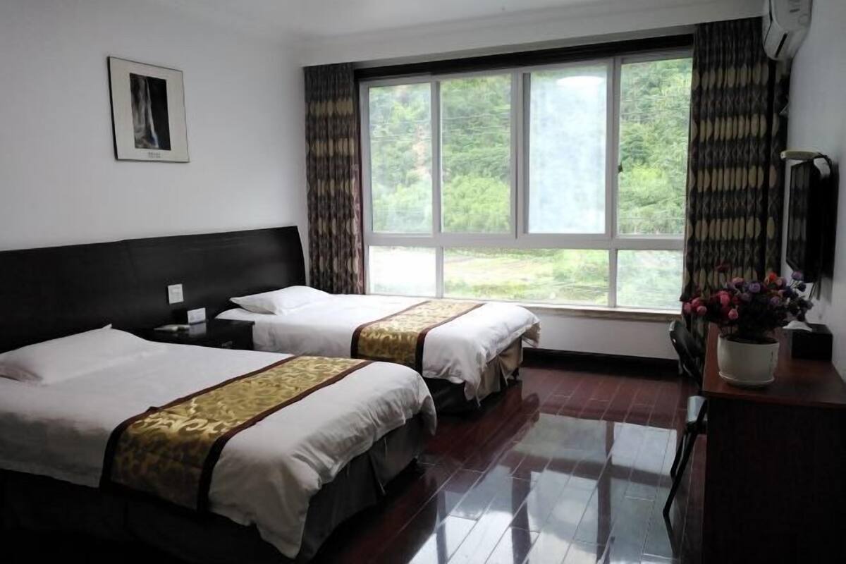 A hotel room with two beds, a large window with curtains, a desk with a vase of flowers, and a framed picture on the wall.