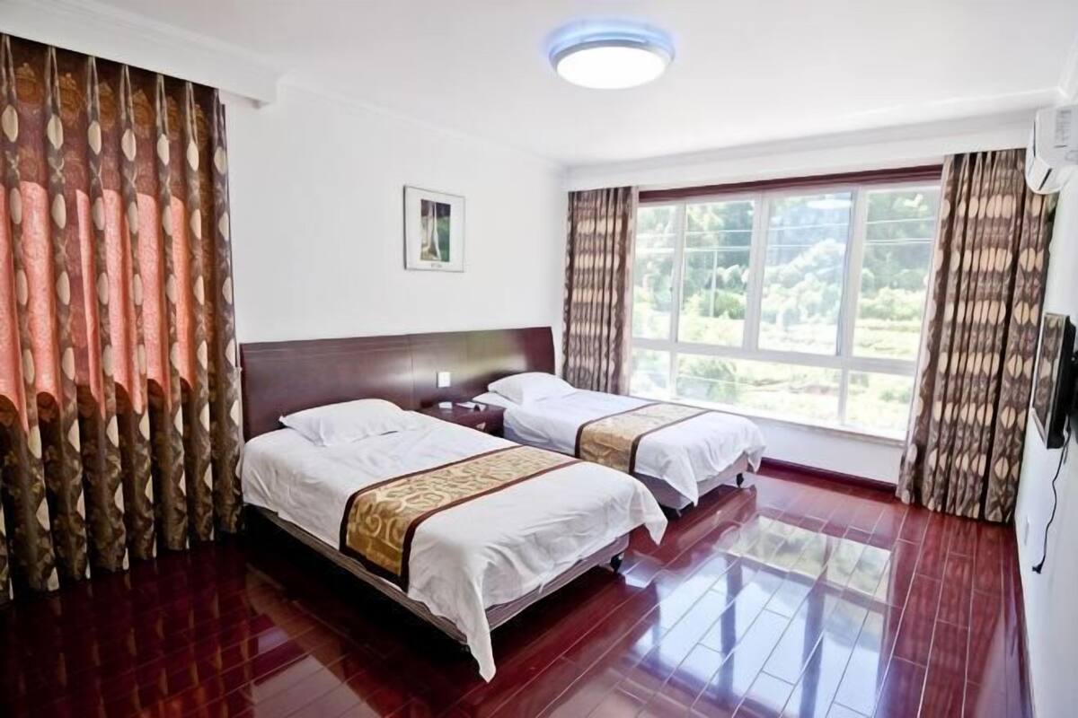 A hotel room with two beds, wooden flooring, a large window, and curtains.