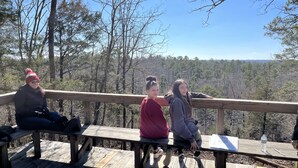 Three people sitting on a wooden bench with a view of a forest. - Entire Campground - All 10 Cabins for Events and Overnights. Up to 40 Guests. (Hatfield)