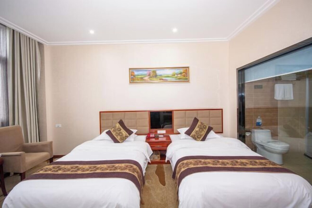 A hotel room with two beds, a TV, a painting, a chair, and a bathroom with a toilet and shower.