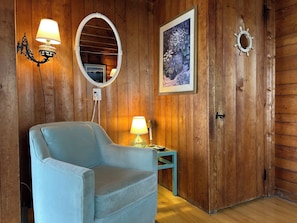 A room with wooden walls, a light blue armchair, two wall-mounted lamps, a framed picture, and a door with a round mirror.