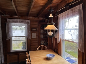 A wooden cabin interior with a dining table, chairs, a hanging lamp, and a view of a lake through large windows.
