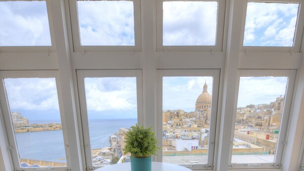 Ein Zimmer mit einem runden Tisch, zwei türkisfarbenen Stühlen, einer Topfpflanze und großen Fenstern mit Blick auf Stadt und Meer.