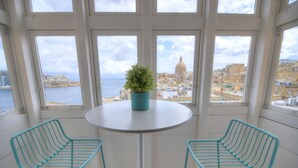 Ein Zimmer mit einem runden Tisch und zwei türkisfarbenen Stühlen, einer Topfpflanze auf dem Tisch sowie einem Blick auf eine Stadtlandschaft und das Meer durch große Fenster.