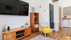 A modern living room with a flat-screen TV, wooden entertainment unit, a yellow armchair, a small potted plant, and a kitchen area with a white dishwasher.