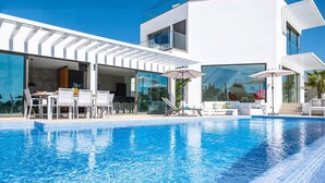 A modern poolside area with a clear blue pool, white outdoor furniture, and a glass-enclosed dining area.