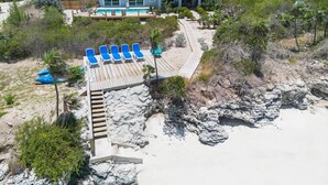 Een strandpand met een groot huis, een houten terras met blauwe ligbedden en een rotsachtige kustlijn.